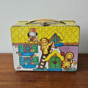 Pode incluir: Lancheira de metal vintage com fundo amarelo e um design extravagante de casa de gengibre. A lancheira apresenta uma alça, fecho e ilustrações de bonecos de gengibre, janelas e flores. Um colecionável charmoso.
