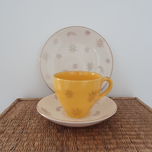 May include: A yellow teacup with a gold star pattern sits on a saucer with a matching pattern. The saucer is on top of a larger plate with a white background and a gold star pattern.