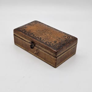 May include: A rectangular brown leather box with a decorative floral pattern on the lid. The box has a clasp closure and gold-coloured stitching around the edges. The box is approximately 15 cm long.