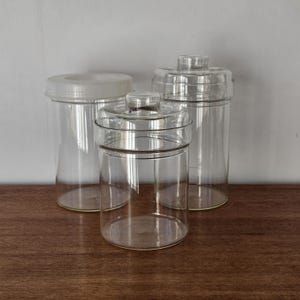 Op de afbeelding: Drie doorzichtige glazen potten met deksels. De potten zijn cilindrisch, één met een wit deksel en de andere twee met doorzichtige glazen deksels. Verschillende maten, geschikt voor de keuken.