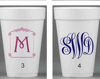 May include: Four white foam cups with different monogram designs. Cup 1 has a black "M" in a decorative frame. Cup 2 has a gold "AMK" in a decorative frame. Cup 3 has a pink "M" in a decorative frame. Cup 4 has a blue "SJD" in a decorative frame.
