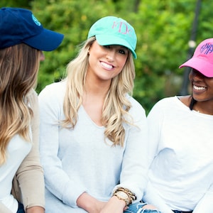 May include: Three women wearing personalized baseball caps. The caps are blue, turquoise, and pink with white embroidered initials. The women are smiling and looking at the camera.