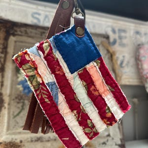 May include: A fabric keychain featuring a quilted American flag design made from various floral and solid red, white, and blue textile patches. The flag is attached to a brown leather strap with a metal snap and a metal swivel lobster clasp.