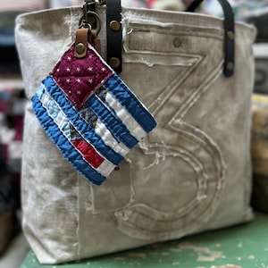 May include: A beige canvas tote bag with black leather handles and a large stitched number three on the front. A quilted fabric charm featuring a red, white, and blue star-patterned design hangs from a brass clip on the handle.