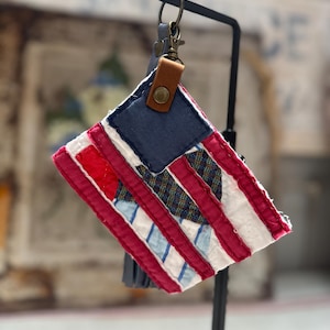 May include: A small, rectangular fabric pouch featuring a patchwork design resembling the American flag. It includes red, white, and blue fabric strips, a dark blue square section, and a brown leather tab with a brass snap, attached to a metal ring.