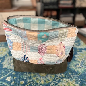 May include: A handmade patchwork zippered pouch with a green canvas base. The top section features a variety of fabric patches in pastel colors and floral patterns. The interior lining is a light blue and white gingham pattern.