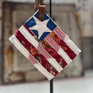 May include: A fabric keychain featuring a rectangular flag design with a blue corner square containing a white star. The flag has alternating white stripes and red floral patterned stripes, attached to a brown leather strap with a metal clasp.