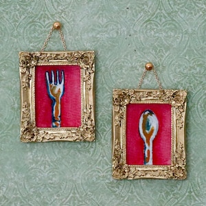 May include: Two framed artworks featuring a fork and spoon design. Each piece has a gold ornate frame, a red background, and a chain for hanging. The fork and spoon are painted in shades of blue, white, and gold.