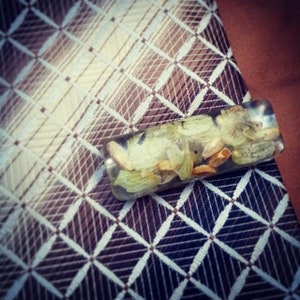 May include: A clear rectangular tie clip containing dried hops and seeds, resting on a patterned tie. The tie features a brown, white, and blue diamond grid design. The tie clip is a unique accessory.