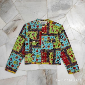 May include: A colorful patchwork denim jacket with a button-up front and long sleeves. The jacket features a variety of heart and star patterns in shades of red, blue, green, and yellow. The label on the jacket reads "VERSACE".