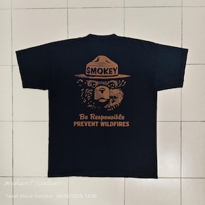 Pu&ograve; includere: T-shirt nero con un'illustrazione marrone di Smokey Bear con un cappello, la parola "SMOKEY" sopra la testa dell'orso. Sotto, il testo recita "Be Responsible PREVENT WILDFIRES".