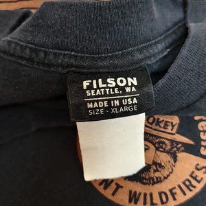 Pu&ograve; includere: Primo piano di una maglietta blu navy scuro con un'etichetta che recita "FILSON SEATTLE, WA MADE IN USA SIZE - XLARGE". La maglietta presenta una grafica di Smokey Bear e le parole "PREVENT WILDFIRES".