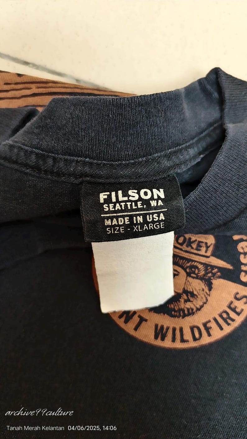 Pu&ograve; includere: Primo piano di una maglietta blu navy scuro con un'etichetta che recita "FILSON SEATTLE, WA MADE IN USA SIZE - XLARGE". La maglietta presenta una grafica di Smokey Bear e le parole "PREVENT WILDFIRES".