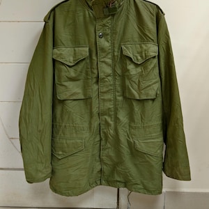 May include: An olive green military-style jacket with multiple front pockets and a high collar. The jacket has a button-up front and shoulder epaulets. The fabric appears to be a durable material, suitable for outdoor wear.
