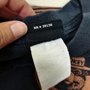Pu&ograve; includere: Primo piano di un'etichetta di una maglietta nera con la scritta "RN # 39126" in bianco. Un'etichetta bianca &egrave; attaccata sotto. La maglietta ha un design grafico con le parole "SMOKEY" e "WILDLIFE".