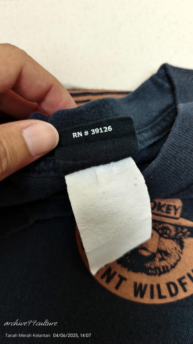 Pu&ograve; includere: Primo piano di un'etichetta di una maglietta nera con la scritta "RN # 39126" in bianco. Un'etichetta bianca &egrave; attaccata sotto. La maglietta ha un design grafico con le parole "SMOKEY" e "WILDLIFE".