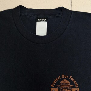 Pu&ograve; includere: T-shirt nera con grafica circolare di Smokey Bear e la scritta "Protect Our Forests, Prevent Wildfires". La maglietta ha scollo tondo e un'etichetta "FILSON SEATTLE, WA".