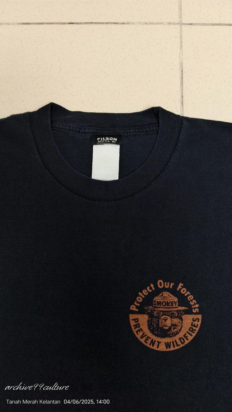 Pu&ograve; includere: T-shirt nera con grafica circolare di Smokey Bear e la scritta "Protect Our Forests, Prevent Wildfires". La maglietta ha scollo tondo e un'etichetta "FILSON SEATTLE, WA".