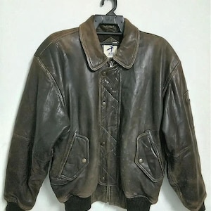 May include: A brown leather bomber jacket with a zipper closure and a ribbed knit collar, cuffs, and waistband. The jacket has a logo patch on the left sleeve and a small pocket on the left chest.
