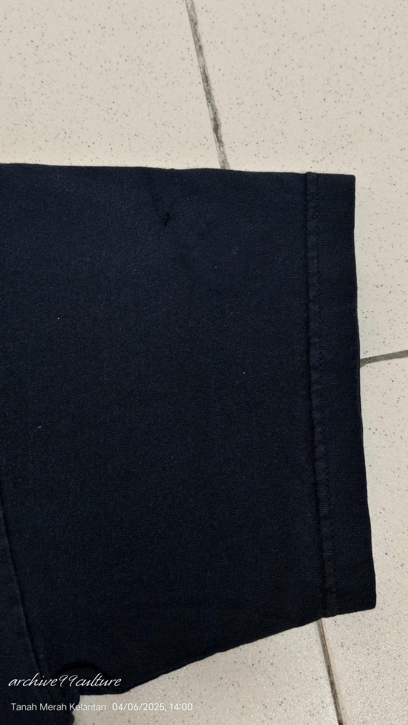 Pu&ograve; includere: Primo piano di un tessuto nero, probabilmente una maglietta o un altro capo di abbigliamento. L'immagine mostra una parte dell'articolo, compresa una cucitura e il bordo del tessuto. Il testo "archive99culture" e le informazioni sulla data sono visibili.