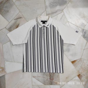 May include: White short-sleeved polo shirt with black and white vertical stripes. The shirt has a collar and a button closure. The Nike logo is on the left chest.