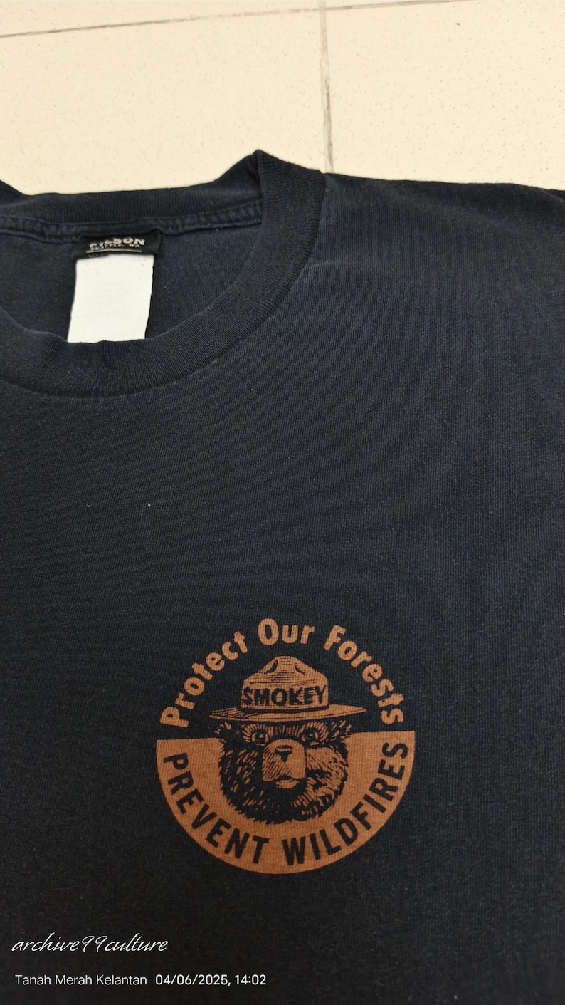 Pu&ograve; includere: T-shirt blu navy scuro con grafica circolare di Smokey Bear con cappello da ranger, con testo "Protect Our Forests" e "Prevent Wildfires". La maglietta ha girocollo e un'etichetta sul retro.