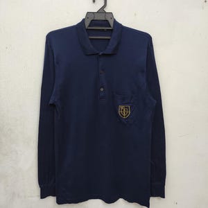 Pode incluir: Polo de manga comprida azul marinho com gola e carcela de três botões. A camisa apresenta um pequeno bolso no peito com um emblema dourado. A camisa está pendurada num cabide preto.