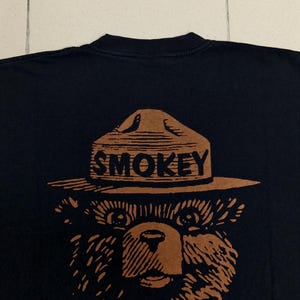 Pu&ograve; includere: T-shirt nera con un'illustrazione marrone di Smokey Bear con cappello da ranger e la scritta "SMOKEY". Sotto c'&egrave; il testo "Be Responsible PREVENT WILDFIRES".