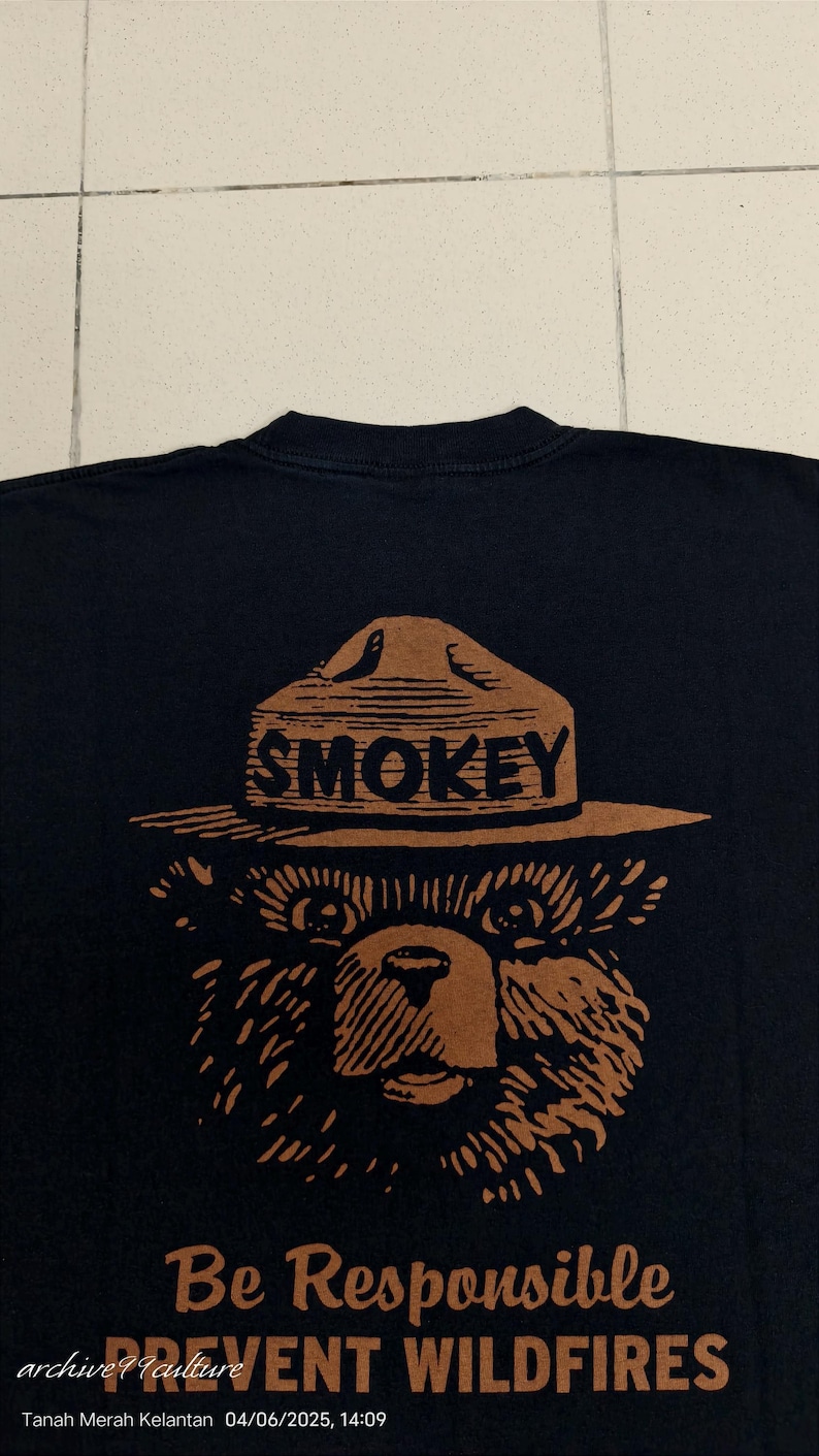 Pu&ograve; includere: T-shirt nera con un'illustrazione marrone di Smokey Bear con cappello da ranger e la scritta "SMOKEY". Sotto c'&egrave; il testo "Be Responsible PREVENT WILDFIRES".