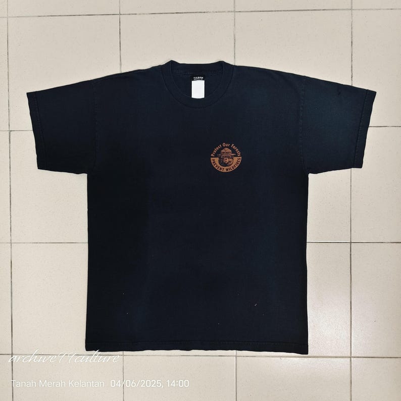 Pu&ograve; includere: T-shirt nera con girocollo e maniche corte. Una grafica circolare sul petto recita "Protect Our Forests, Prevent Wildfires" in un design marrone e arancione. La maglietta &egrave; piatta.