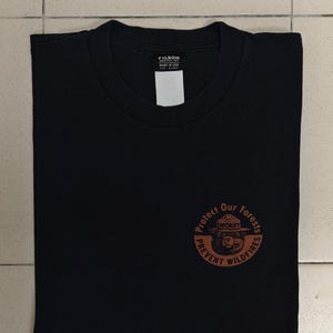 Pu&ograve; includere: T-shirt nero con scollo tondo e grafica circolare sul davanti. La grafica presenta Smokey Bear e il testo "Protect Our Forests, Prevent Wildfires". La maglietta ha un'etichetta con la scritta "FILSON".