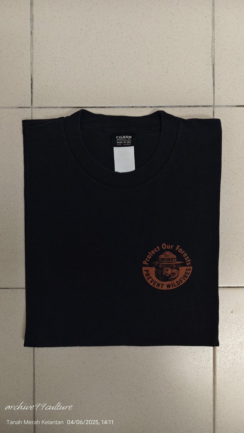 Pu&ograve; includere: T-shirt nero con scollo tondo e grafica circolare sul davanti. La grafica presenta Smokey Bear e il testo "Protect Our Forests, Prevent Wildfires". La maglietta ha un'etichetta con la scritta "FILSON".