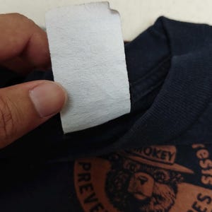 Pu&ograve; includere: T-shirt blu navy con grafica di Smokey Bear e la scritta "PREVENT WILDFIRES". &Egrave; visibile un'etichetta bianca. L'immagine include anche il testo "archive99culture" e la data.