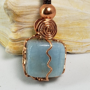 May include: A light blue aquamarine gemstone pendant wrapped in copper wire with a copper bead at the top. The pendant is hanging from a brown cord.
