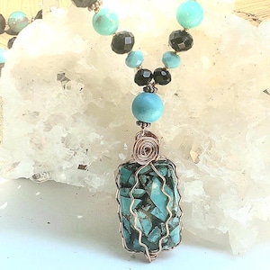 Puede incluir: Un collar artesanal con cuentas turquesas, negras y celeste. El colgante es una piedra rectangular de turquesa envuelta en alambre de cobre. El collar tiene un estilo bohemio único.