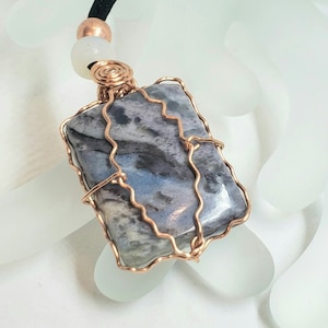 Puede incluir: Colgante rectangular con una piedra azul y gris, envuelta en alambre de cobre. Cuelga de un cordón negro, con una cuenta de cobre y una cuenta blanca. La piedra tiene un aspecto veteado.