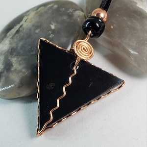 Puede incluir: Colgante triangular negro con envoltura de alambre de cobre, cuentas negras y una espiral de cobre. El colgante está en un cordón negro y descansa sobre una piedra gris y blanca.