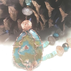 Op de afbeelding: Een handgemaakte ketting met een grote, druppelvormige hanger. De hanger heeft blauwe, groene en gele patronen, omwikkeld met koperdraad. De ketting bevat turquoise en stenen kralen, met een spiraalaccent van koperdraad.
