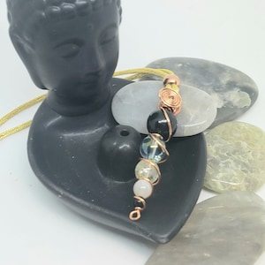 Puede incluir: Una cabeza de Buda de cerámica negra con un cordón amarillo y un collar de cristal con cuentas negras, blancas y azules. El collar está envuelto alrededor de una piedra de cristal blanca.
