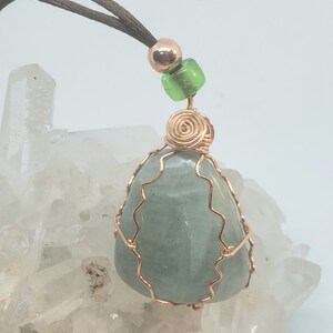 May include: A light green aquamarine gemstone wrapped in copper wire, with a copper wire spiral at the top, and a green glass bead and a copper bead on a brown cord.