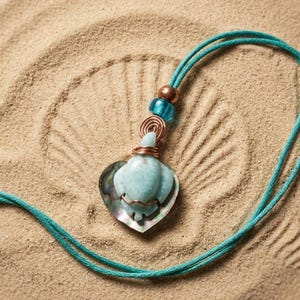 May include: A turquoise necklace with a heart-shaped pendant. The pendant features a light blue turtle design set on an iridescent shell. The necklace has a teal cord with copper and blue bead accents, set against a sandy background.