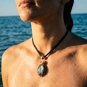 Può includere: Una collana con pendente e cordino nero. Il pendente è una pietra ovale blu-verde, avvolta in filo di rame. La collana è indossata sulla pelle di una persona, con l'oceano sullo sfondo.