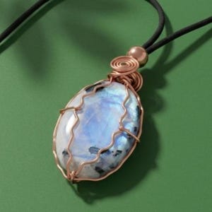 Peut inclure: Un collier pendentif artisanal avec une pierre de lune ovale. La pierre présente des tons blancs, bleus et noirs, sertie dans un cadre en fil de cuivre. Le collier a un cordon noir et un fermoir couleur cuivre.