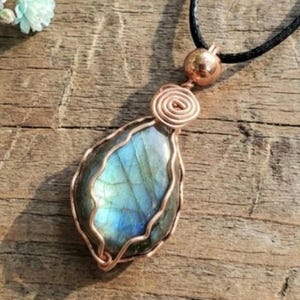 Op de afbeelding: Een ketting met een druppelvormige hanger met een blauwgroene edelsteen, omhuld door koperdraad. De draad is rond de steen gewikkeld en vormt een spiraal. De ketting heeft een zwart koord en een koperkleurige kraal.