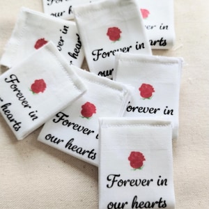 May include: White fabric squares with the text "Forever in our hearts" in black script and a red rose illustration. The squares are arranged in a pile, with several visible.