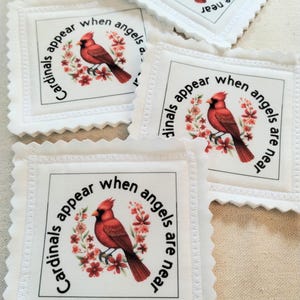 May include: Several white fabric squares with a scalloped edge. Each square features a red cardinal bird illustration surrounded by a floral wreath and the text "Cardinals appear when angels are near."
