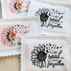 May include: White fabric squares with a scalloped edge feature sunflower designs and the text "A beautiful soul is never forgotten." Some sunflowers are in color, others in black and white, with butterfly accents.