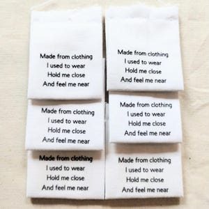 May include: Five rectangular white fabric labels arranged in two columns. Each label features black printed text that reads: Made from clothing, I used to wear, Hold me close, And feel me near. The labels have a clean, woven texture and straight edges.