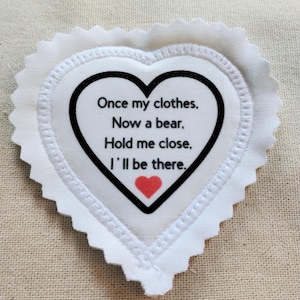 May include: A white heart-shaped fabric patch with pinked edges. The center features a black heart outline containing the text: Once my clothes, Now a bear, Hold me close, I'll be there, followed by a small red heart at the bottom.