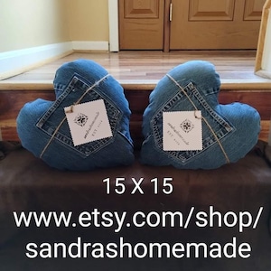 May include: Two heart-shaped pillows crafted from blue denim fabric, each featuring a visible back pocket and a square white tag tied with twine. The text 15 X 15 and www.etsy.com/shop/sandrashomemade is printed in white below the pillows.